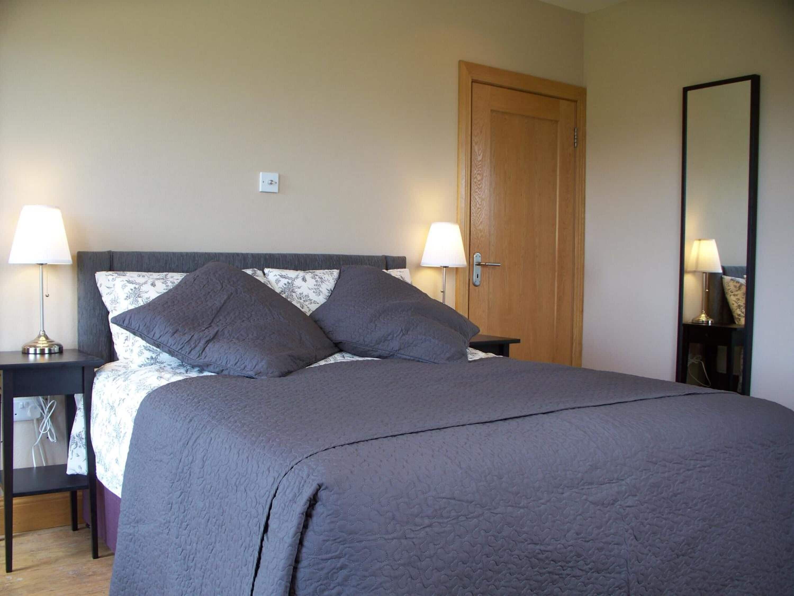 A neatly made bed with a grey duvet and pillows, two bedside tables with lamps, a wooden door, and a mirror on the wall.