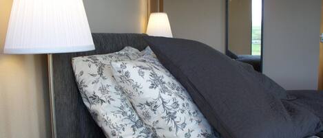 Un lit bien fait, agrémenté d’oreillers à motifs floraux et d’une couverture gris foncé, une lampe de chevet et un miroir au mur.