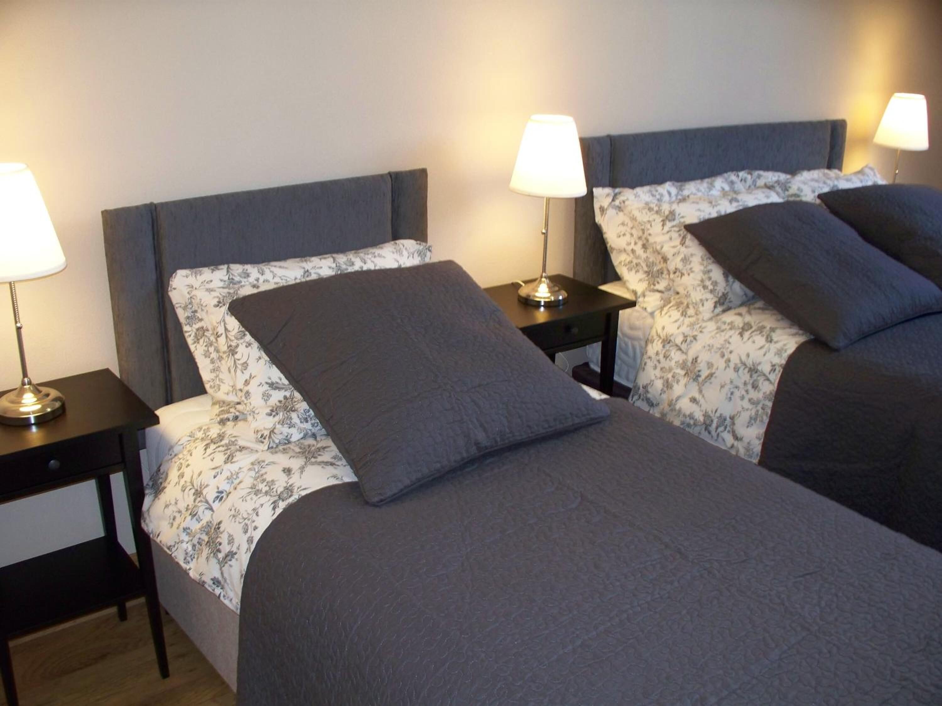 A hotel room with two beds, each with a dark headboard and floral-patterned bedding. There are bedside lamps on each nightstand.