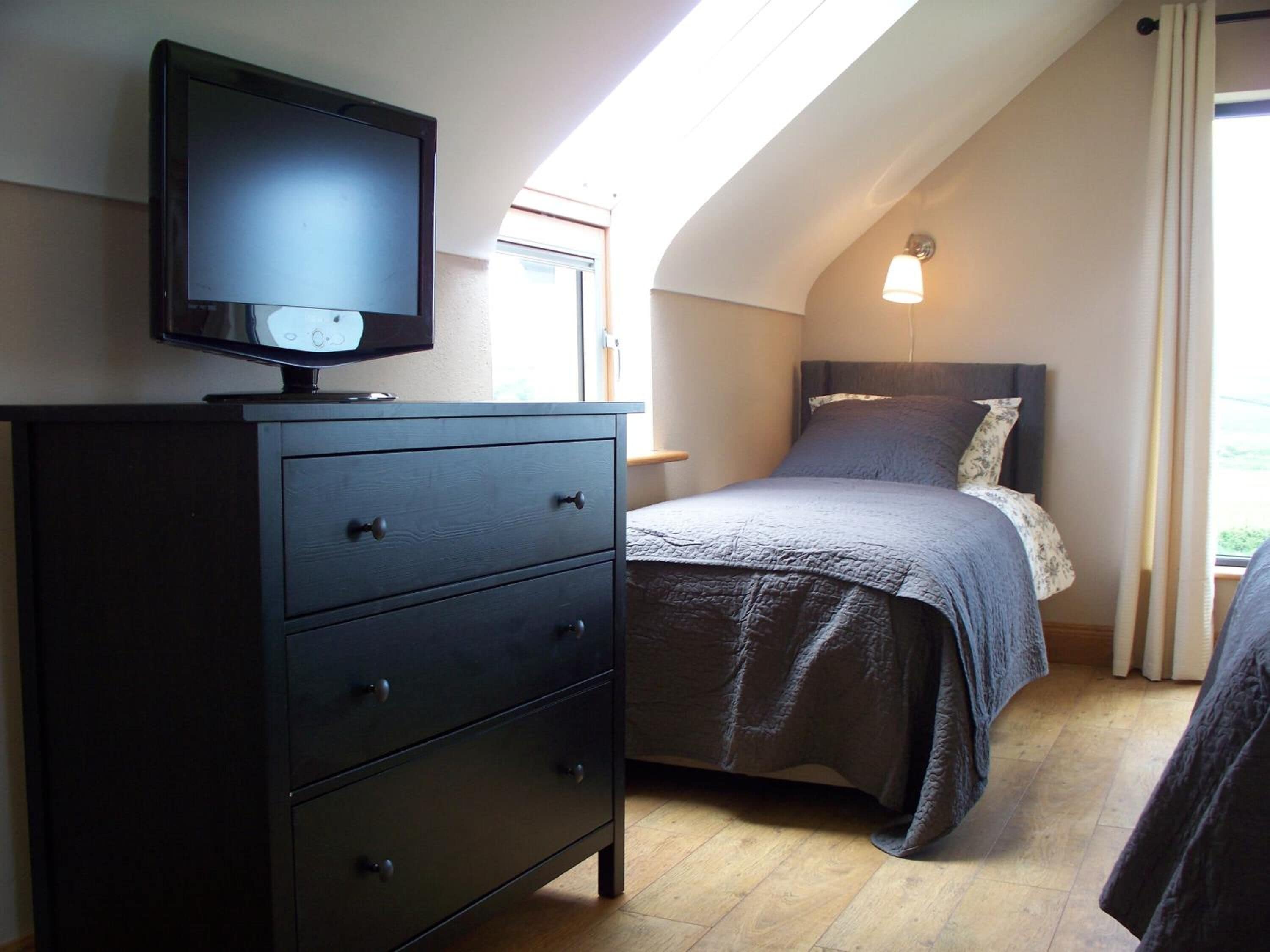 A bedroom with a bed, a dresser, a television on top of the dresser, and a skylight above the bed.