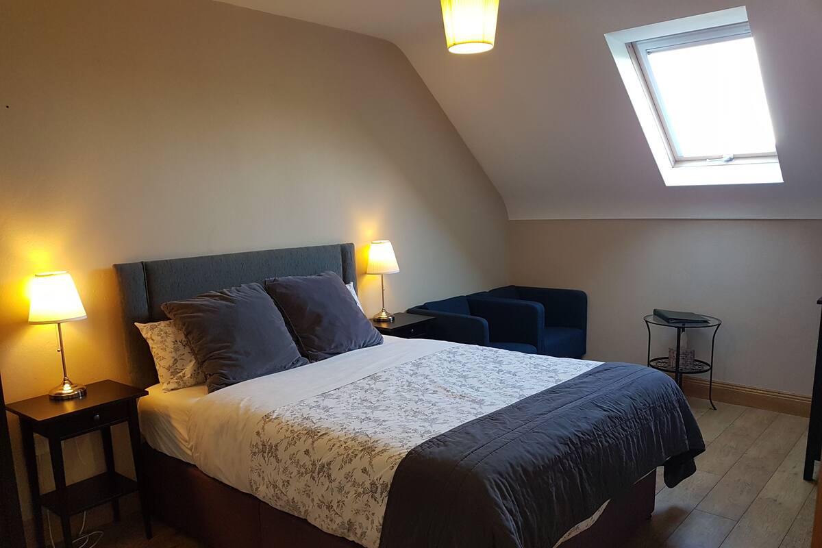 A bedroom with a bed, two bedside lamps, a small table, a chair, and a skylight.