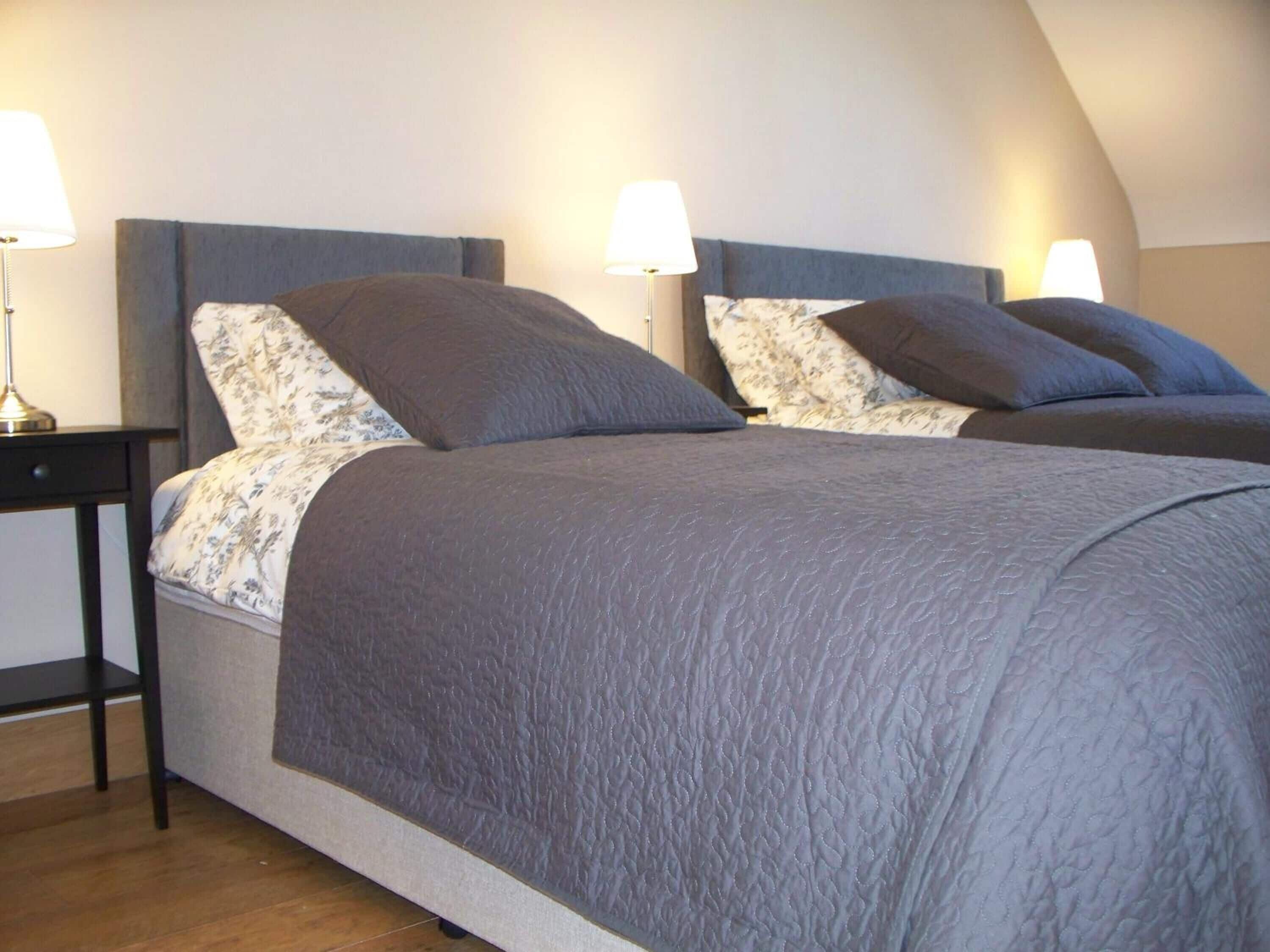 Una cama doble con colcha gris y almohadas estampadas con motivos florales, dos lámparas de noche y una mesita de noche de madera.