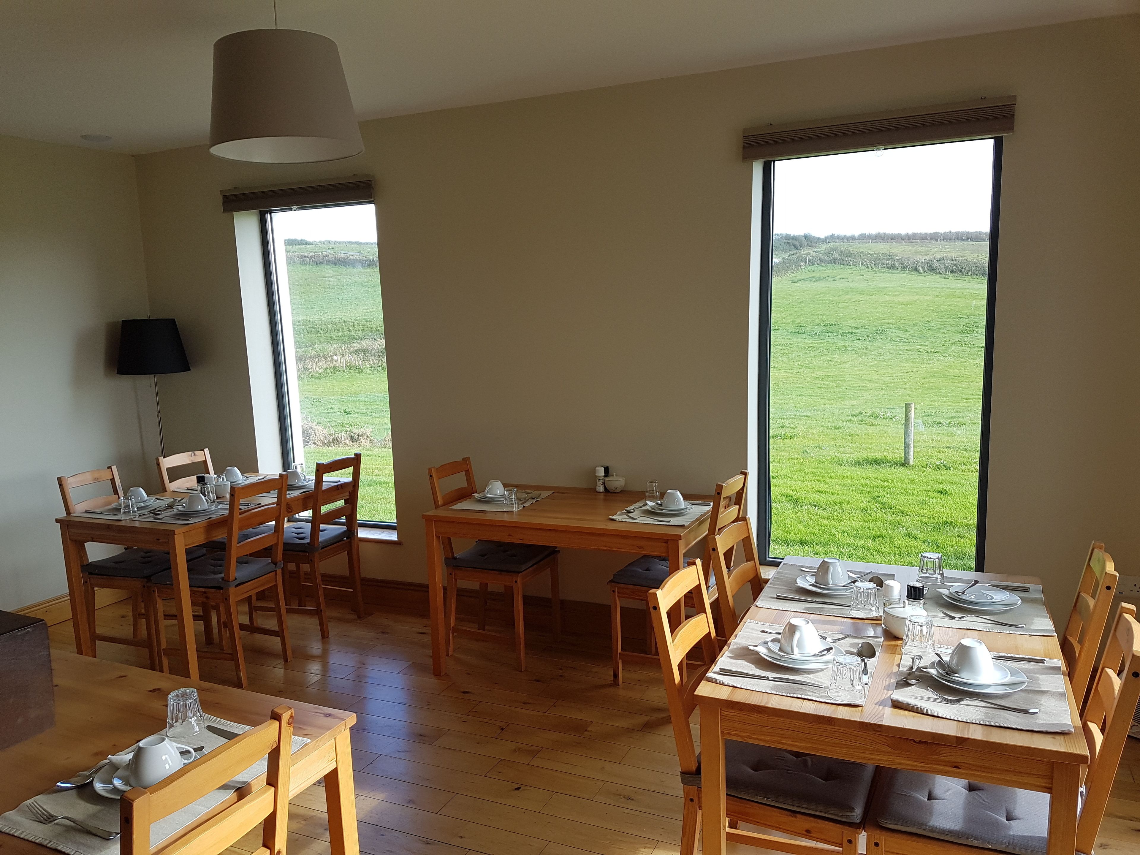 A dining area with wooden tables and chairs, large windows with a view of a green field, and a hanging lamp.