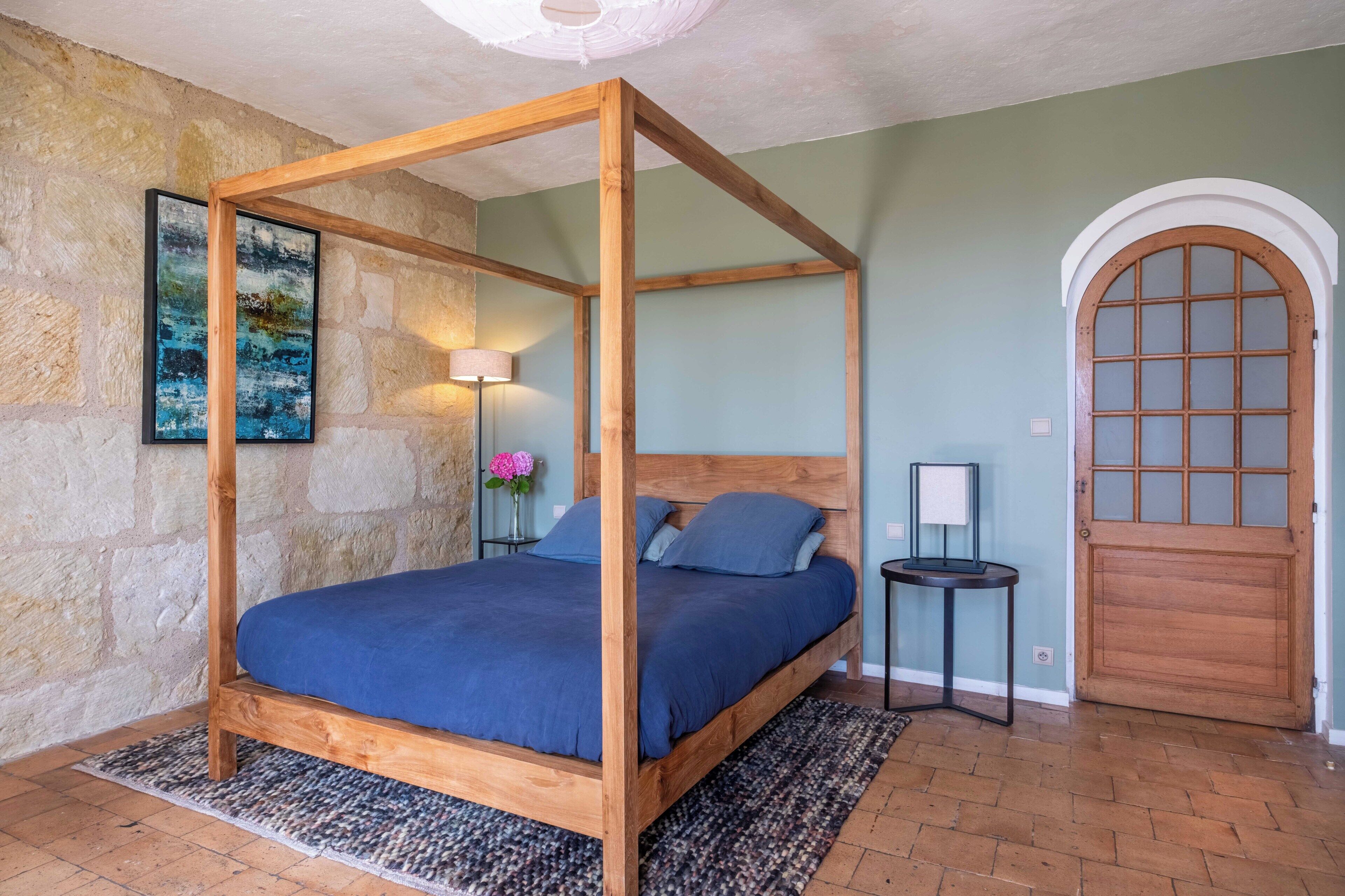 Uma cama de dossel com colcha azul, uma parede de pedra, uma porta de madeira e um quadro pendurado na parede.