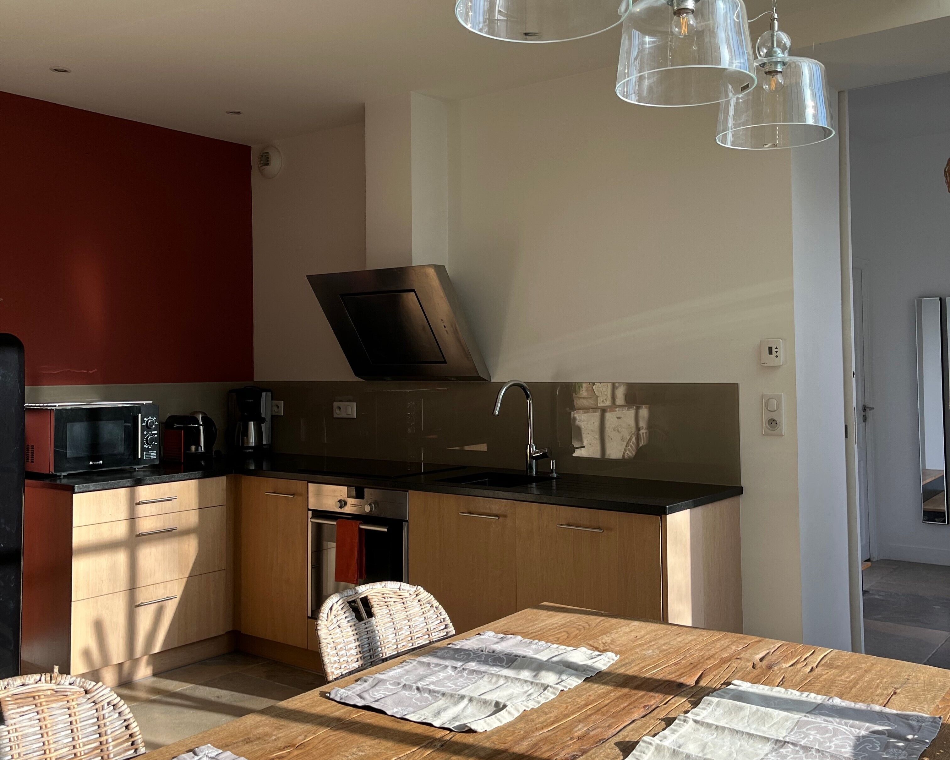 Ett modernt kök med mikrovågsugn, kaffebryggare och en inbyggd ugn. Här finns ett matbord med en bordsduk och en stol av rotting.
