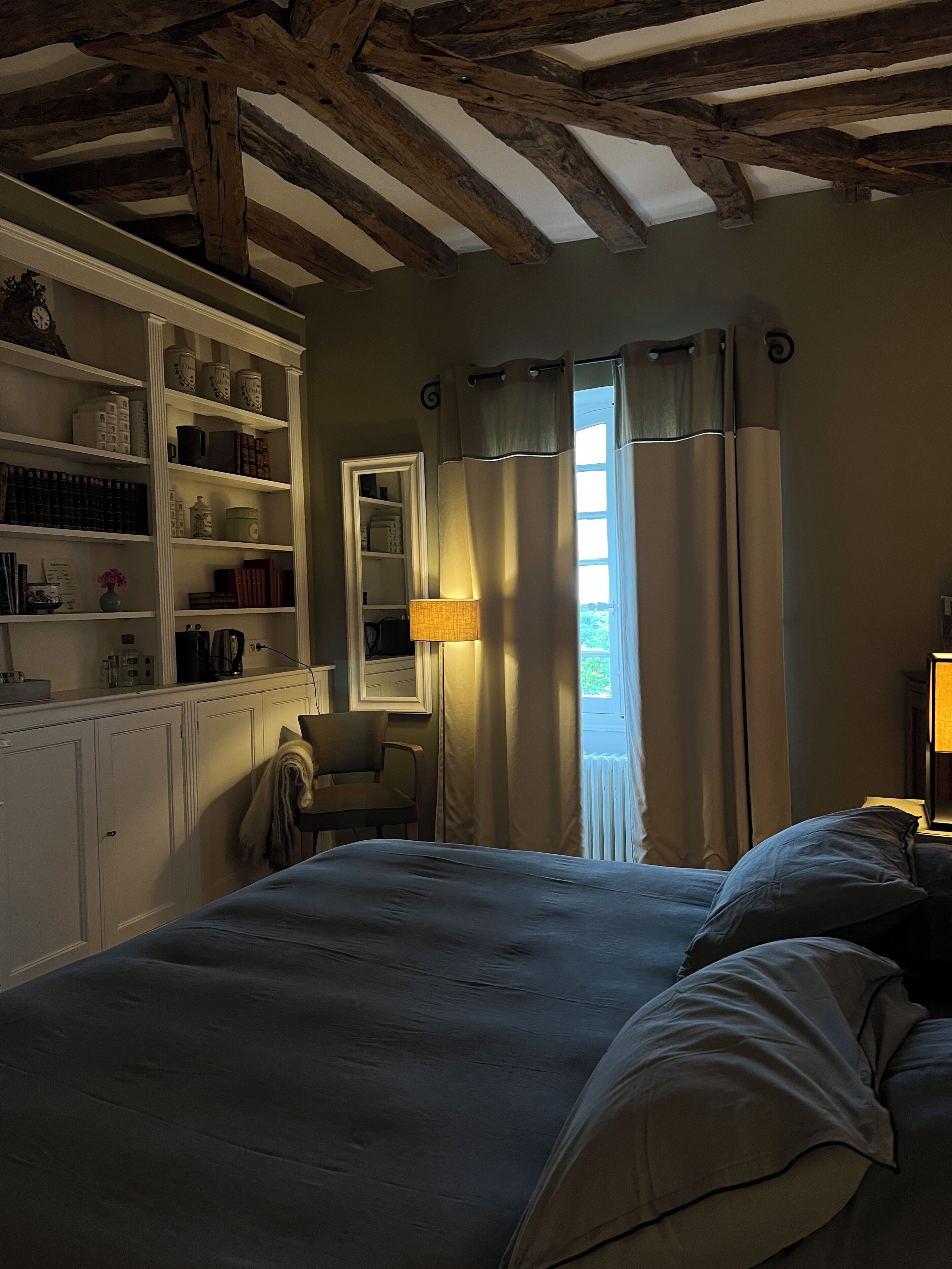 A bedroom with a large bed, a wardrobe, a desk with shelves, a window with curtains, and a lamp.