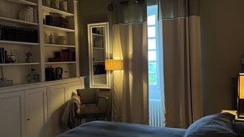 A bedroom with a large bed, a wardrobe, a desk with shelves, a window with curtains, and a lamp.