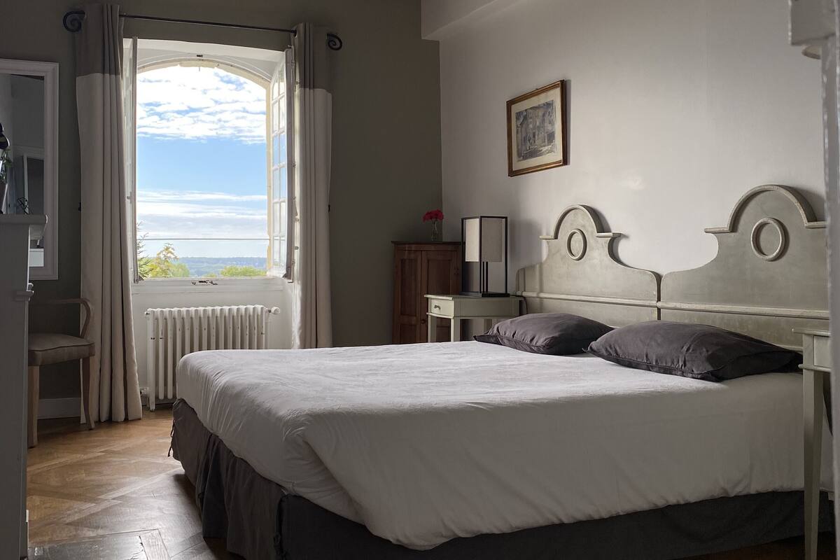 Une chambre à coucher avec un lit, une fenêtre avec des rideaux, un radiateur, une chaise en bois, une table de chevet avec une lampe et un tableau encadré au mur.