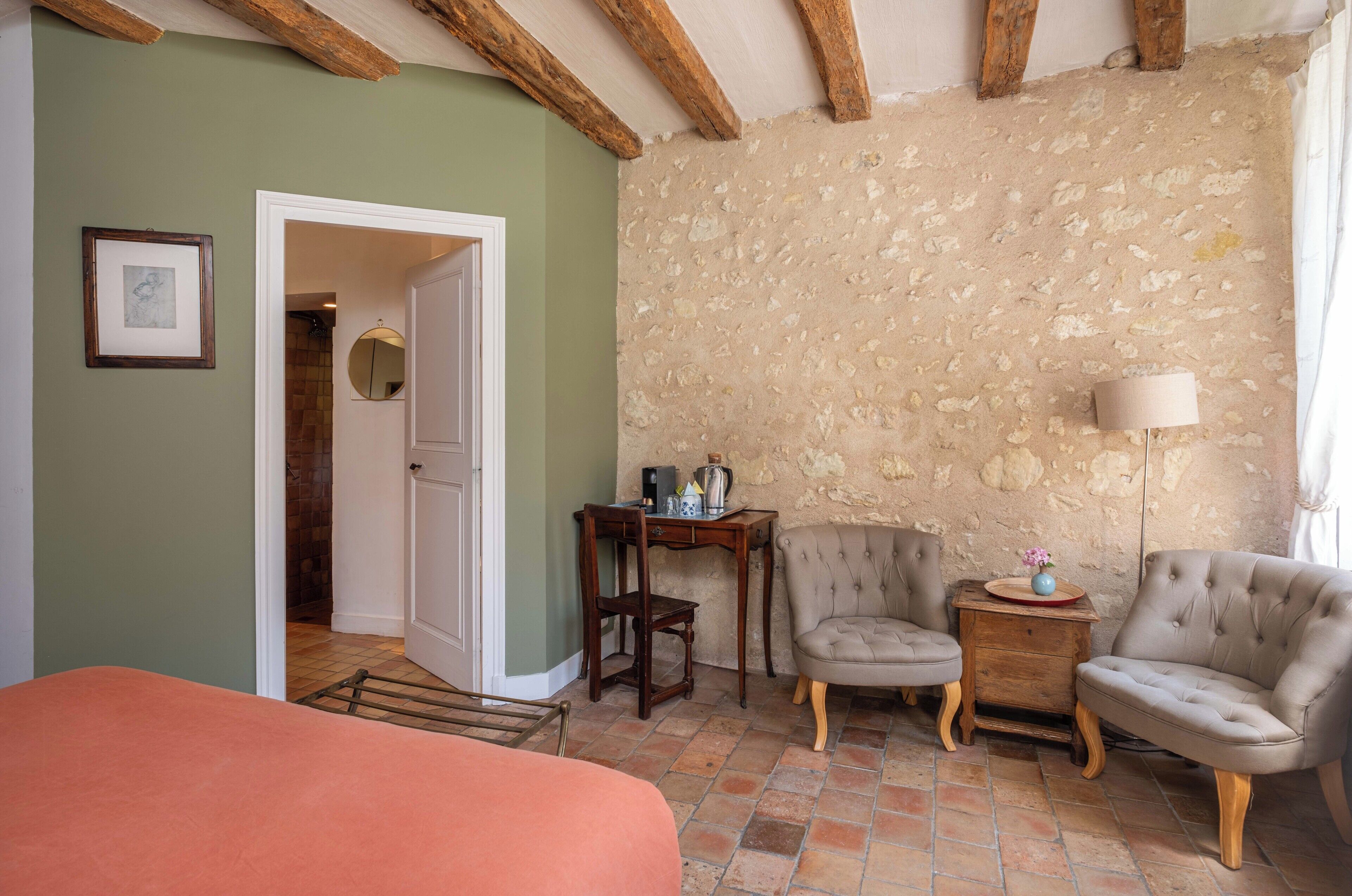 A room with a bed, a wooden table, two armchairs, a lamp, and a stone wall.