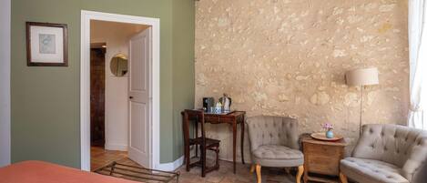 A room with a bed, a wooden table, two armchairs, a lamp, and a stone wall.