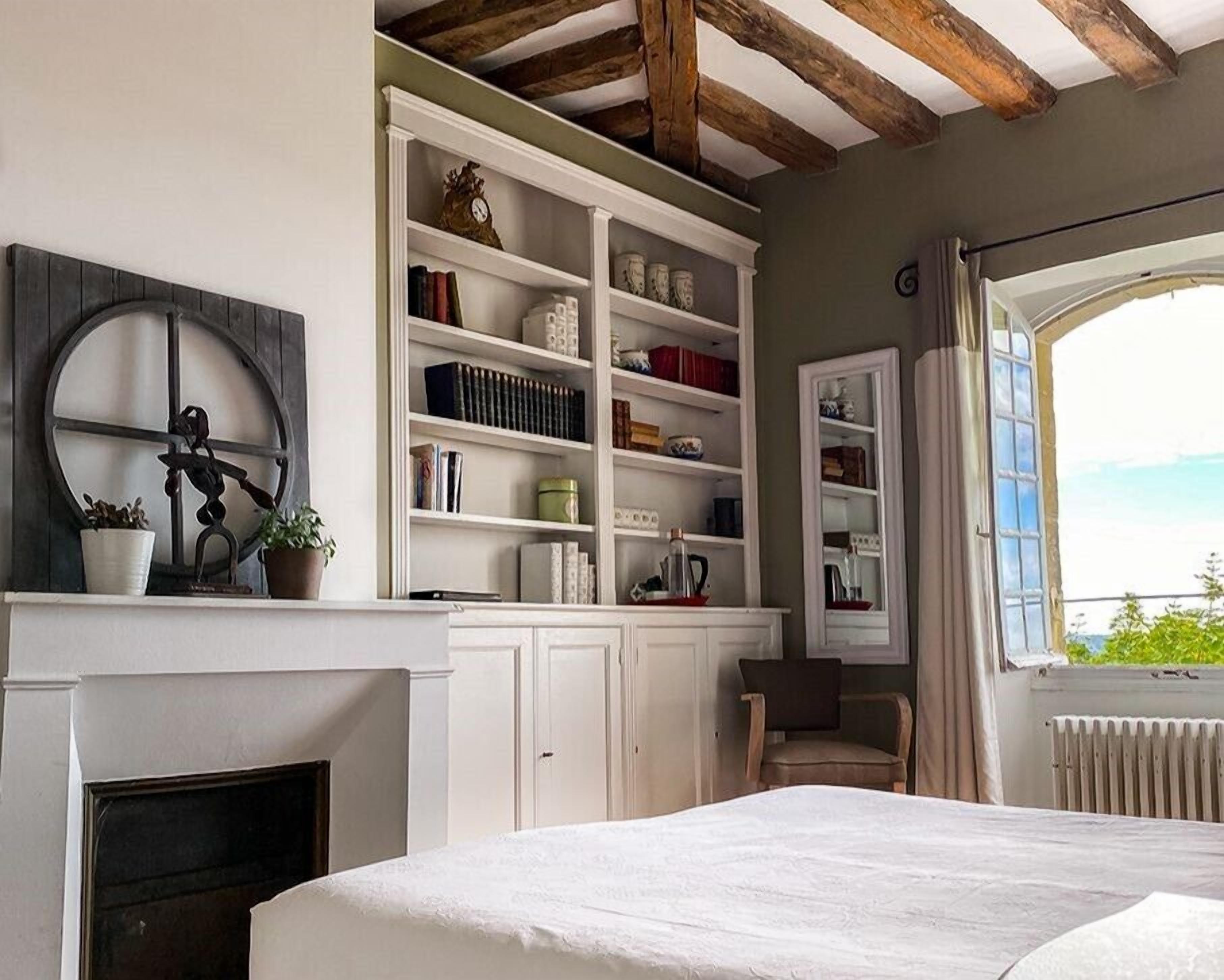 A bedroom with a fireplace, built-in shelving, a bed, and a view through a window.