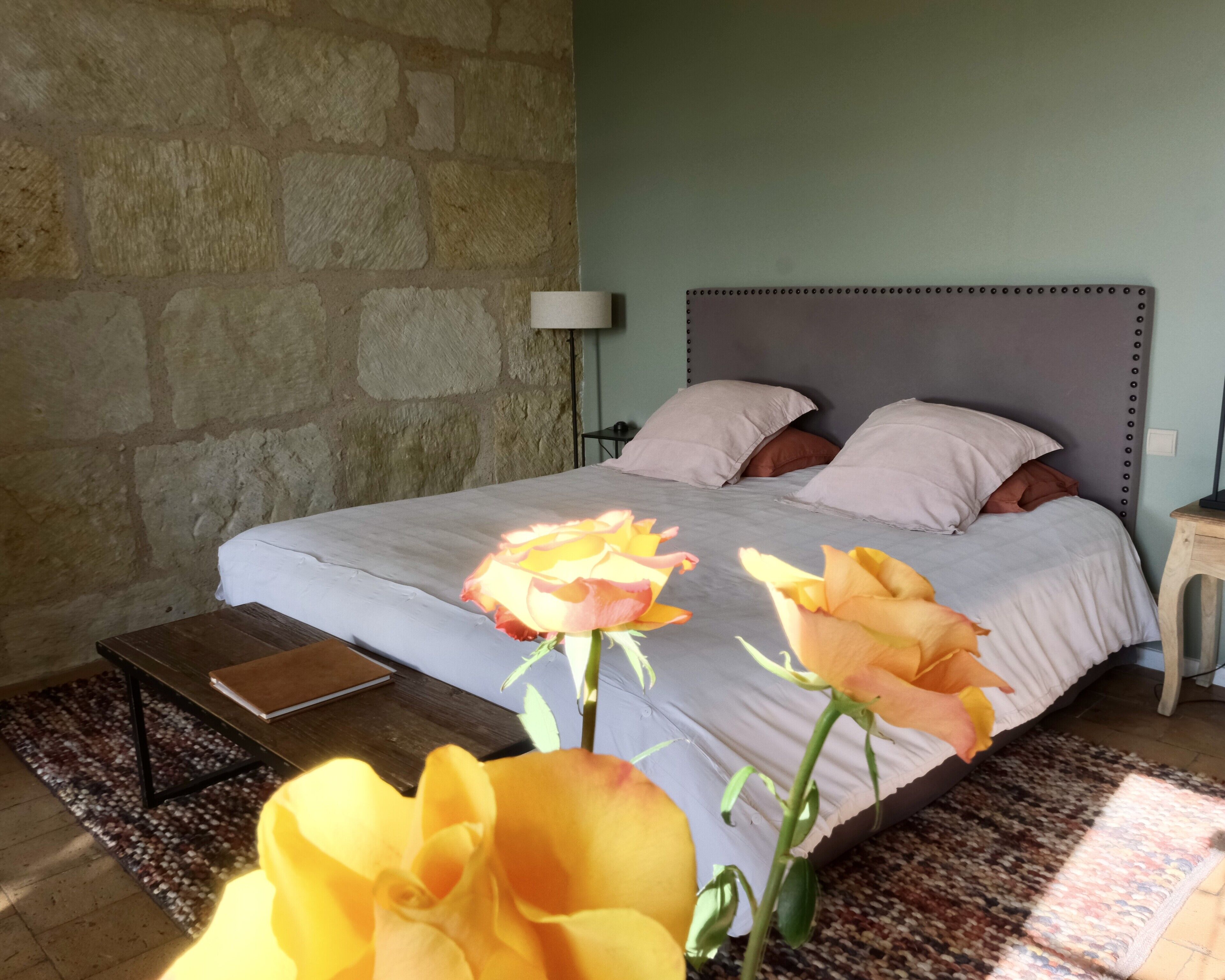 A bedroom with a stone wall, a bed with pillows, a bedside table, and a lamp.