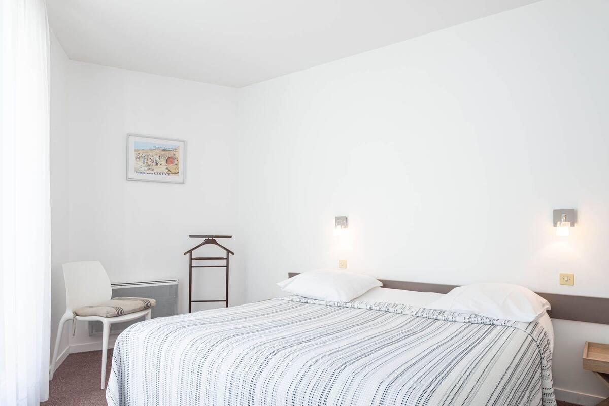A minimalist hotel room with a bed, a chair, a coat rack, and a wall-mounted light fixture.