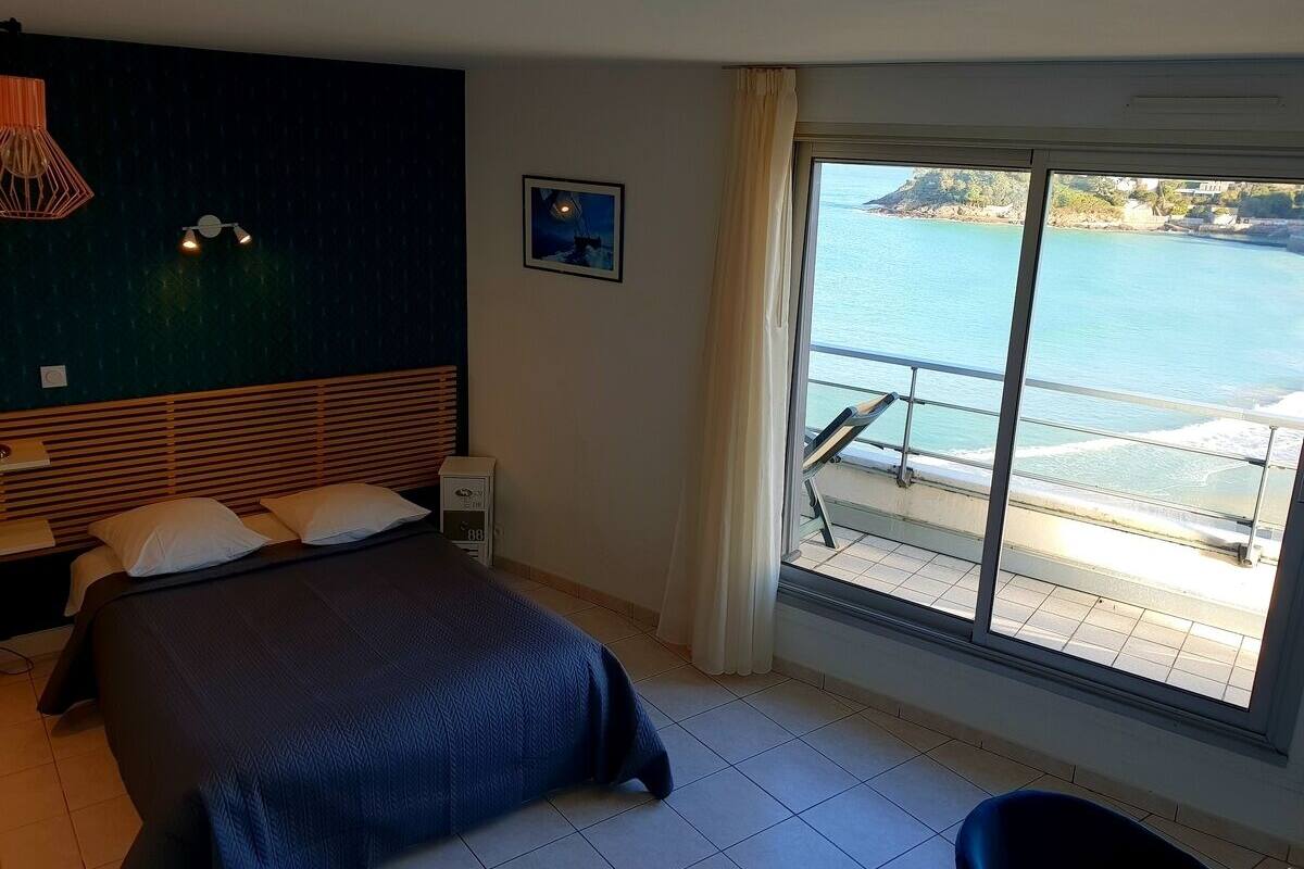 A hotel room with a bed, a bedside table, a nightstand, a wall-mounted lamp, a picture, and a large window with a view of the sea.