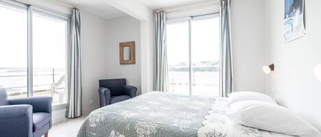 A bedroom with a large bed, a chair, and a view of the sea through large windows.