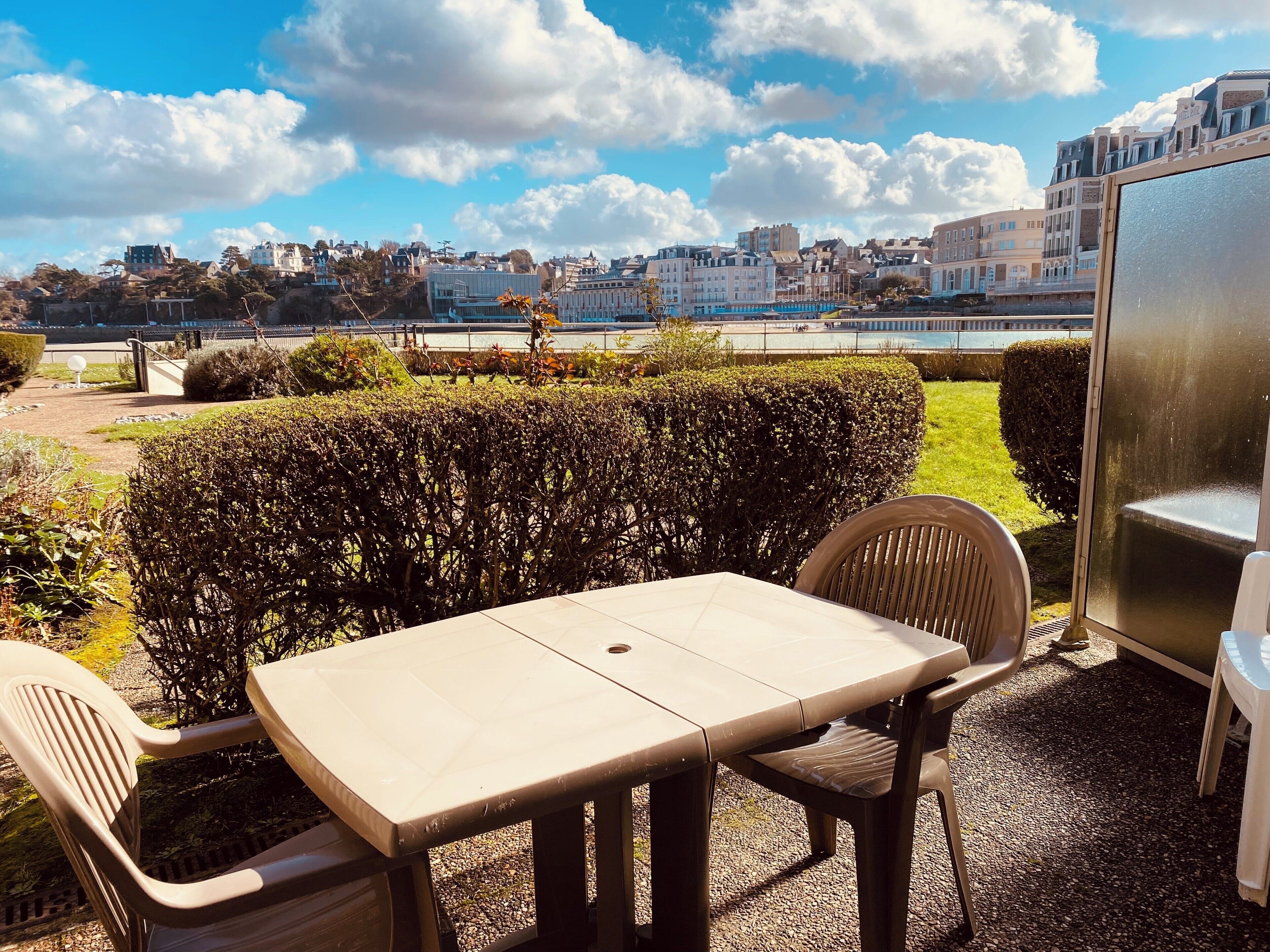 Une terrasse avec une table et des chaises, une haie et une vue sur des bâtiments de l’autre côté d’une étendue d’eau.
