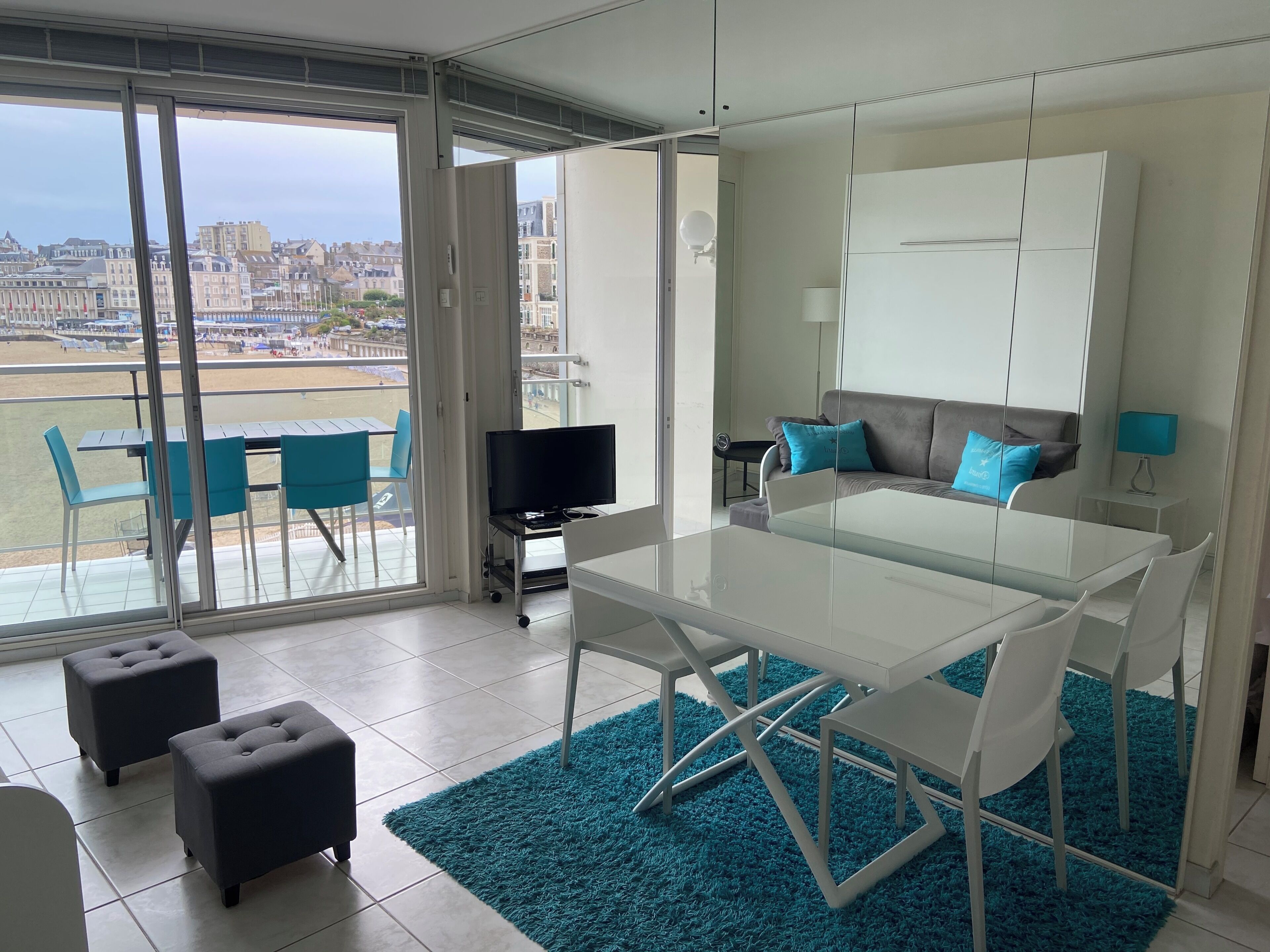 Un salon moderne avec une table en verre, des chaises blanches, un canapé avec des coussins bleus et un téléviseur posé sur un meuble.
