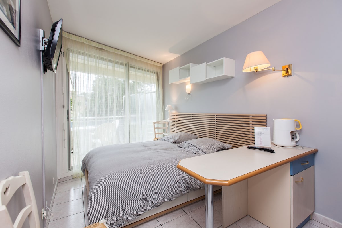 A modern bedroom with a bed, bedside table, lamp, and a view of the outdoors through a window.