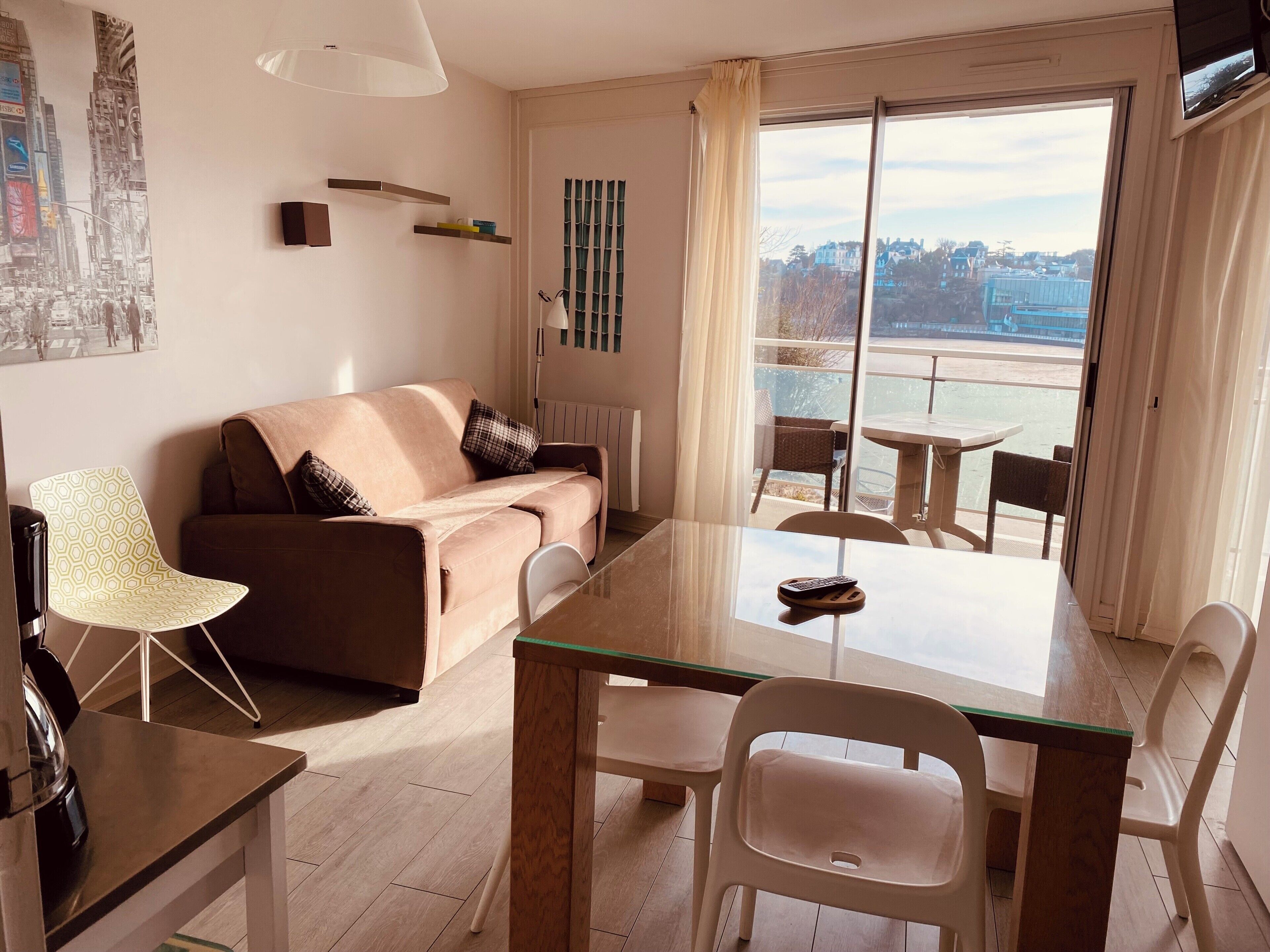 A modern living room with a sofa, a dining table, chairs, a radiator, a TV, and a city view through the window.