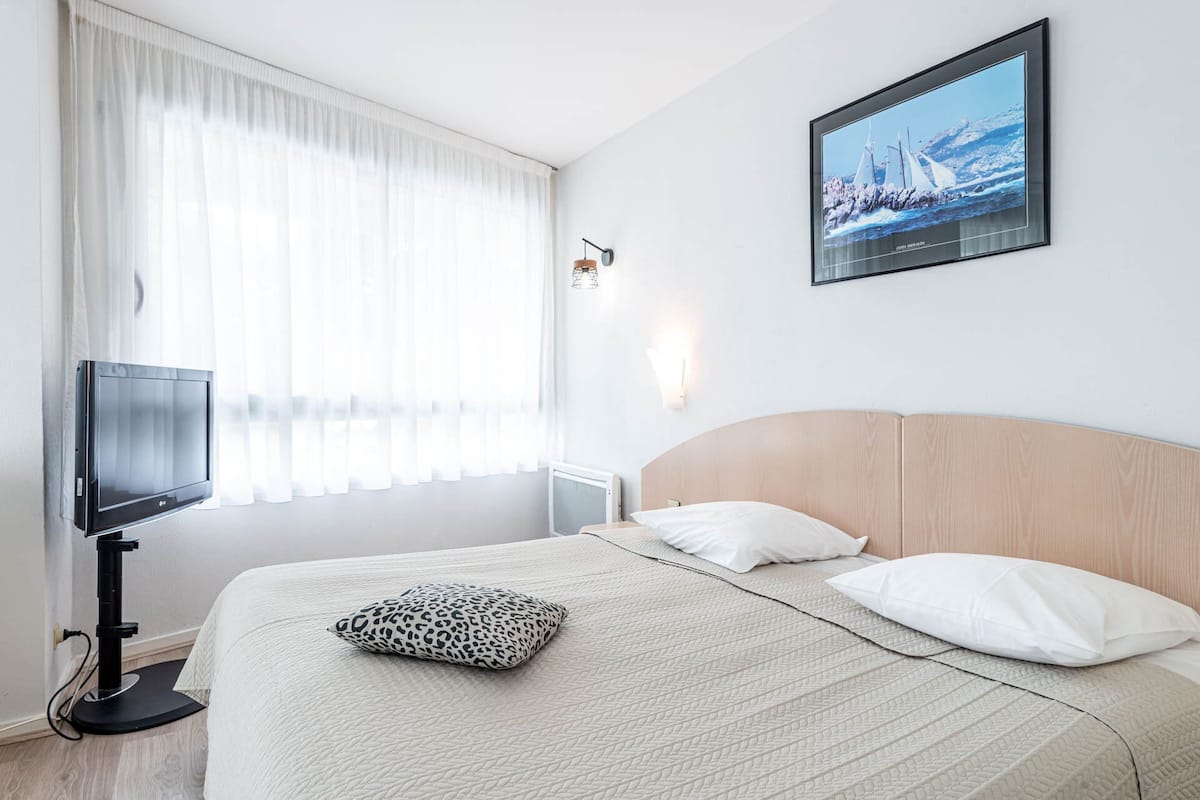 Una habitación de hotel con cama, televisión, ventana con cortinas, un cuadro colgado en la pared y una lámpara de pared.