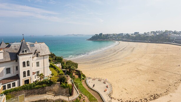 A coastal view with a sandy beach, a promenade with benches, and a historic building with a tower. - Résidence Le Crystal (Dinard)