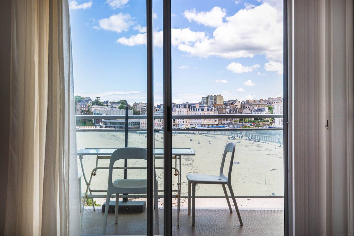Un balcón con una mesa y dos sillas, con vista a una playa y un paisaje urbano con edificios y un puente.