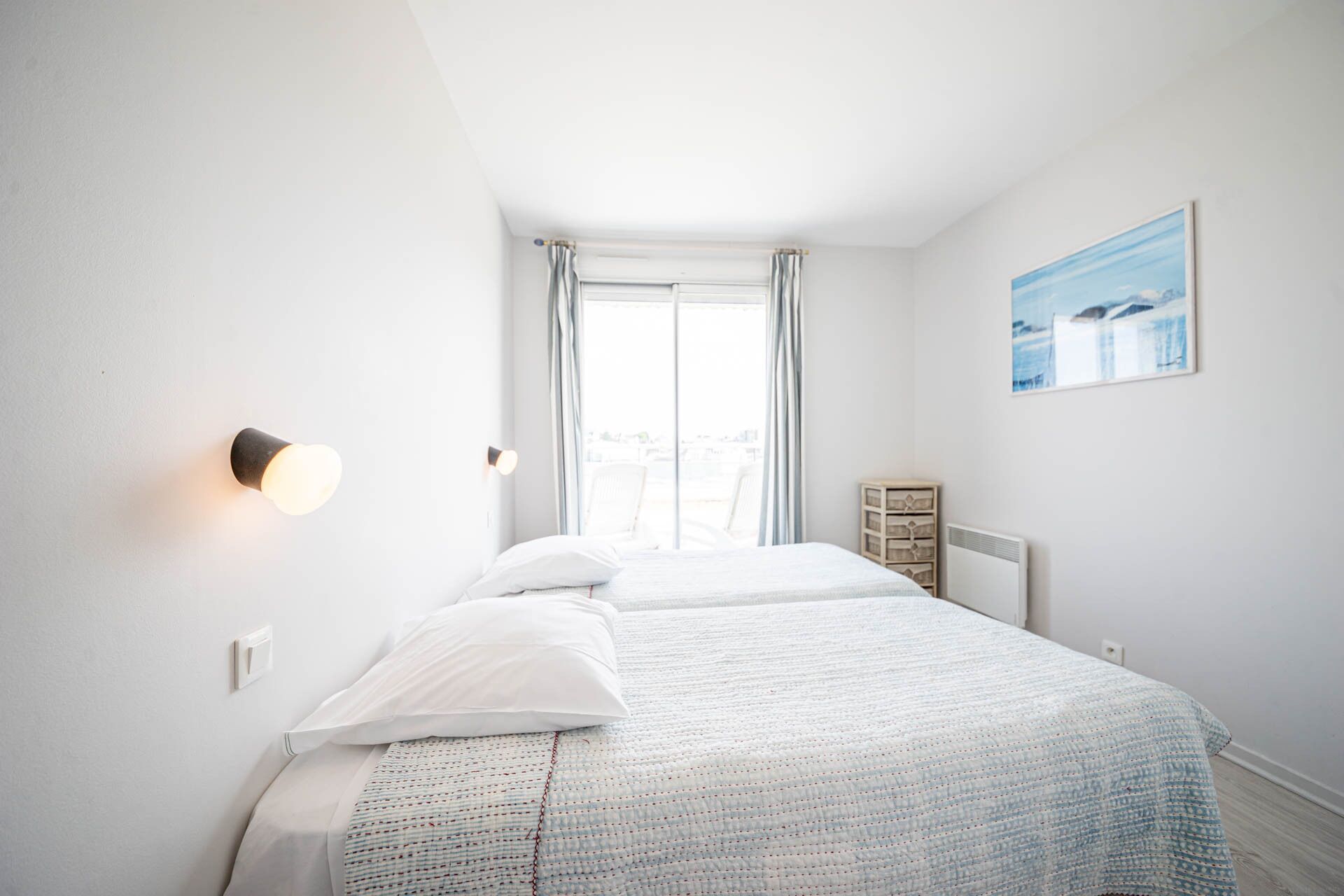A bedroom with two beds, a window with curtains, a wall-mounted light, and a painting.