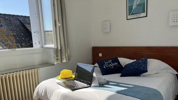 Una cama bien hecha con una computadora portátil, un sombrero amarillo y almohadones decorativos.
