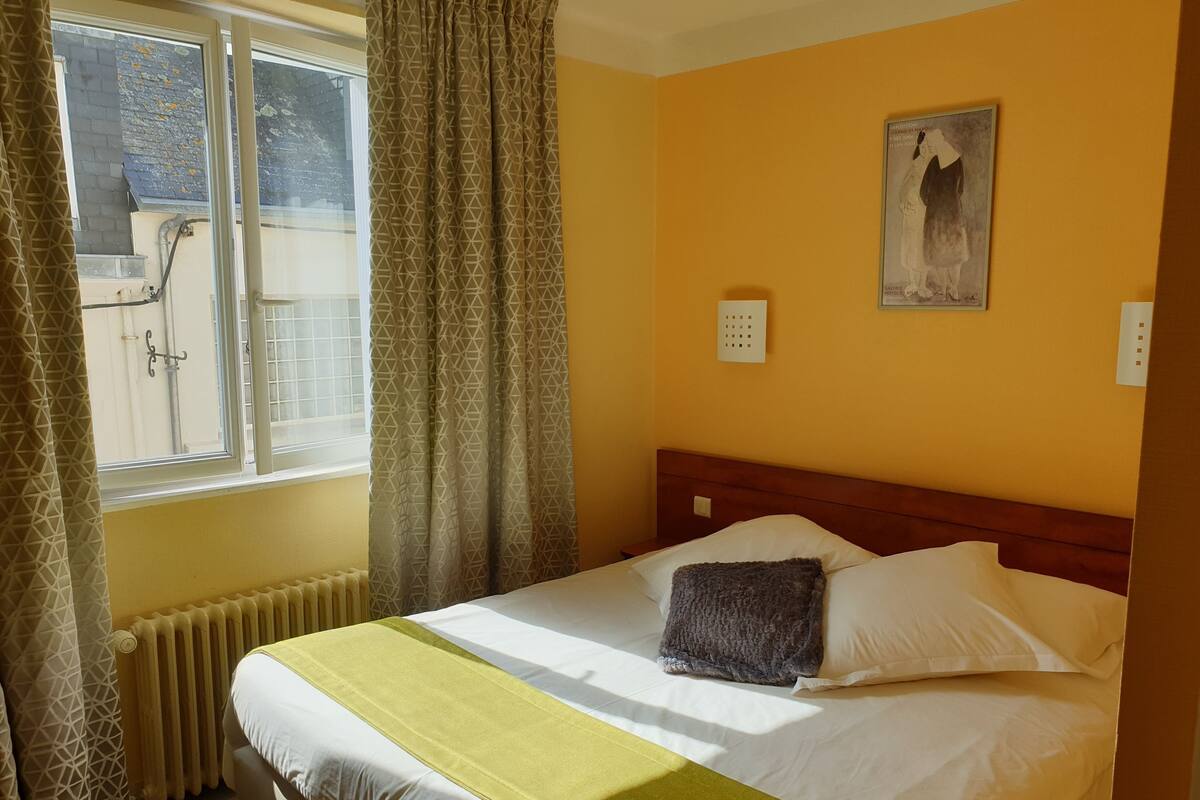 A hotel room with a bed, a radiator, a window with curtains, and a wall-mounted light switch.