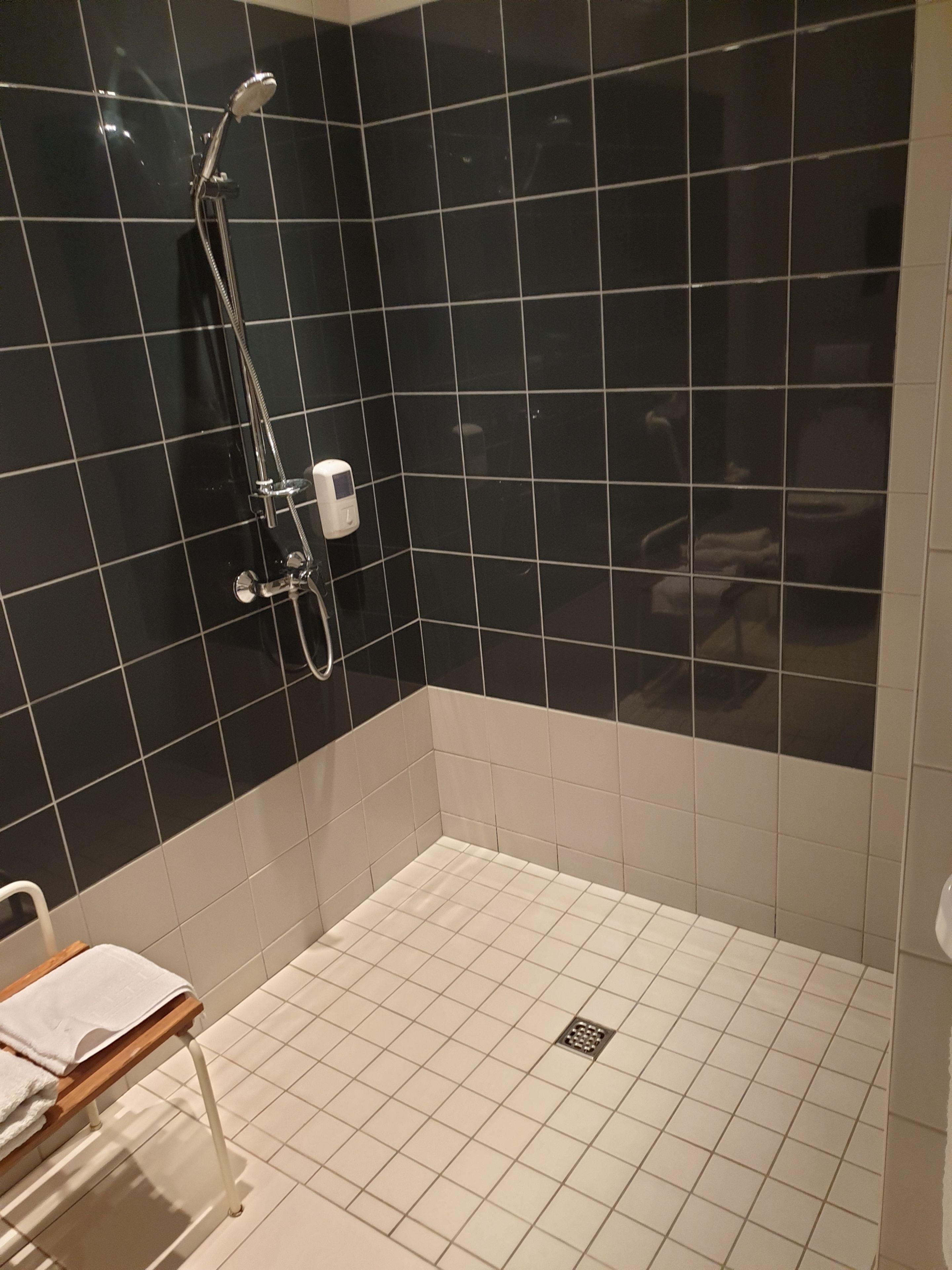 Une salle de bain avec des carreaux noirs et blancs, une douche équipée d’un pommeau de douche amovible et un banc avec des serviettes.