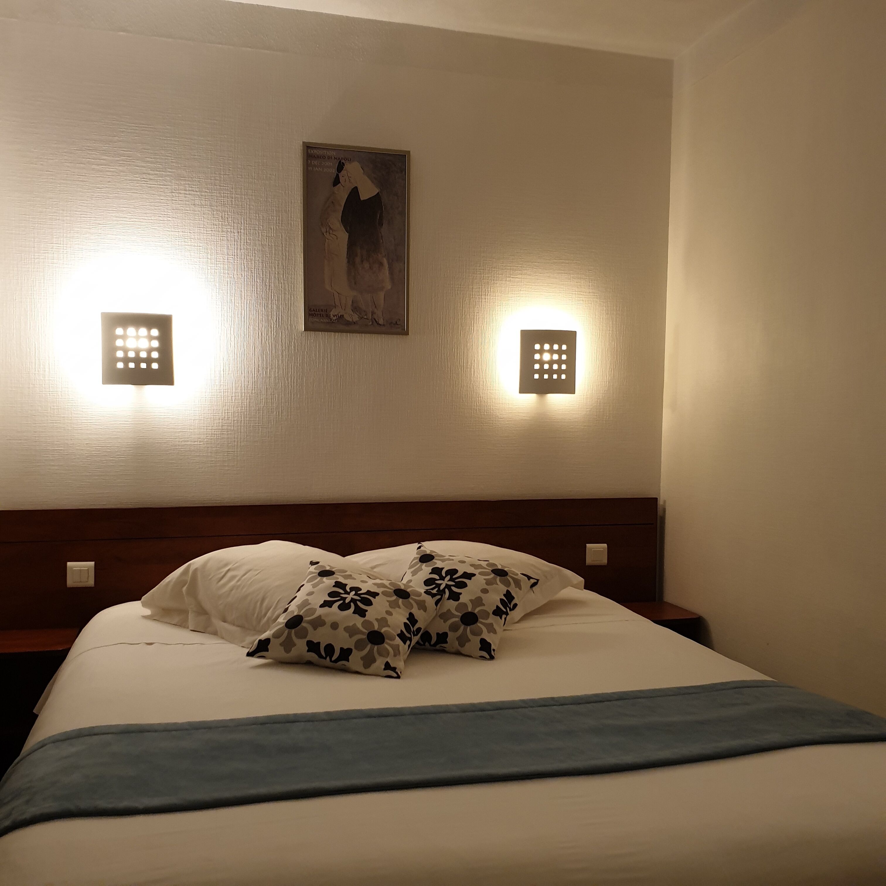 A hotel room with a bed, two wall-mounted light fixtures, and a framed picture on the wall.