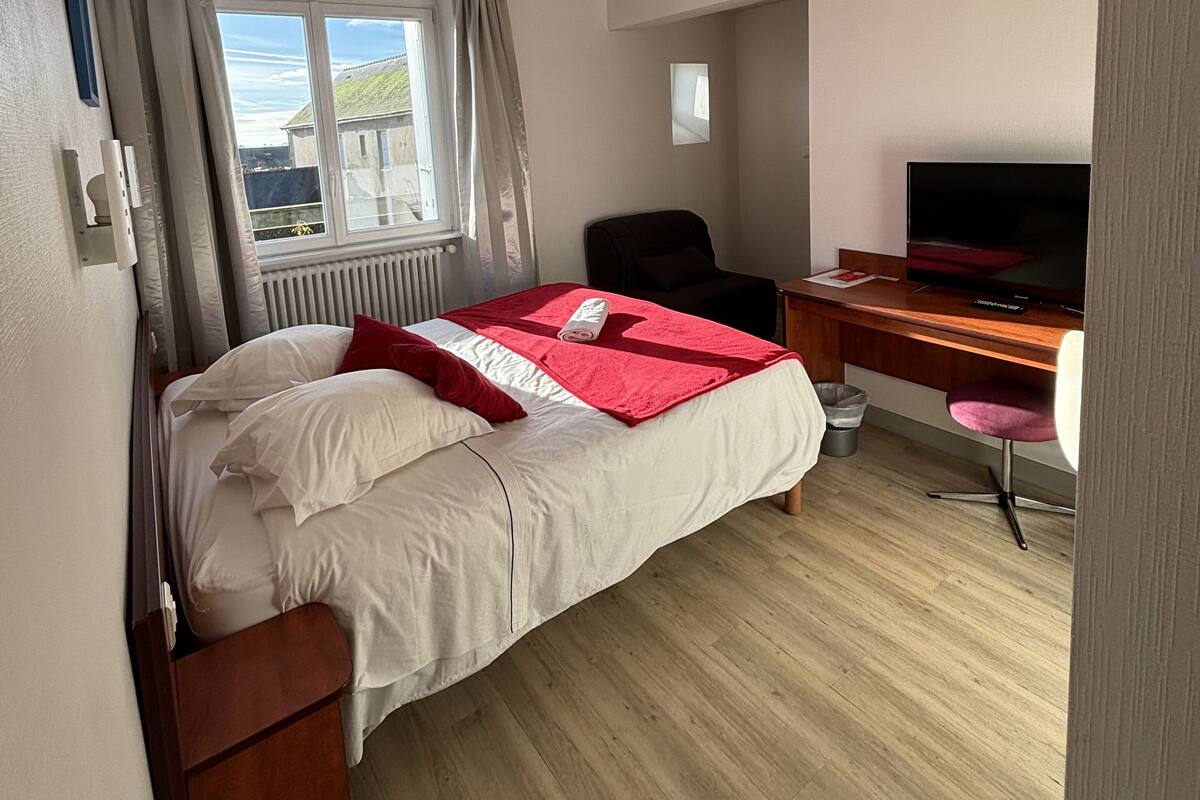 A hotel room with a bed, a desk with a computer, a chair, a TV, and a window with curtains.