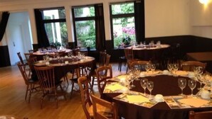 Un comedor con mesas redondas dispuestas para una comida, ventanas amplias con cortinas y vistas al exterior con vegetación.
