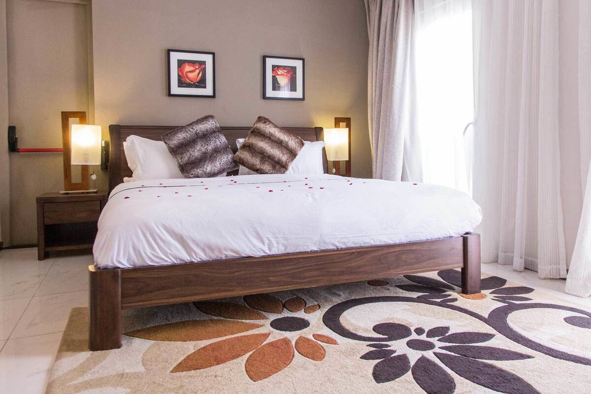 Ein ordentlich bezogenes Bett mit weißer Tagesdecke und zwei Zierkissen, ein Holzkopfstück, zwei Nachttischlampen und ein floral gemustertes Teppich auf Fliesen.