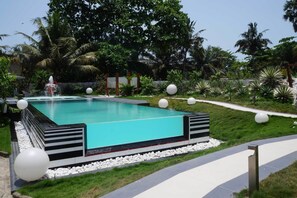 Ein rechteckiger Swimmingpool mit einer Springbrunnenanlage, umgeben von einem gepflegten Garten mit Palmen und weißen, kugelförmigen Dekorationen.