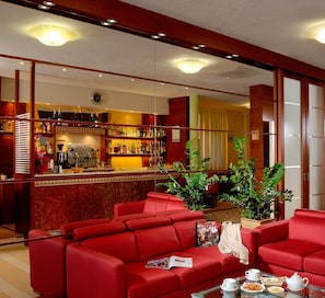Un hall de hotel con sofás rojos, una zona de bar con diversas botellas de licor y un espejo grande que refleja el espacio.