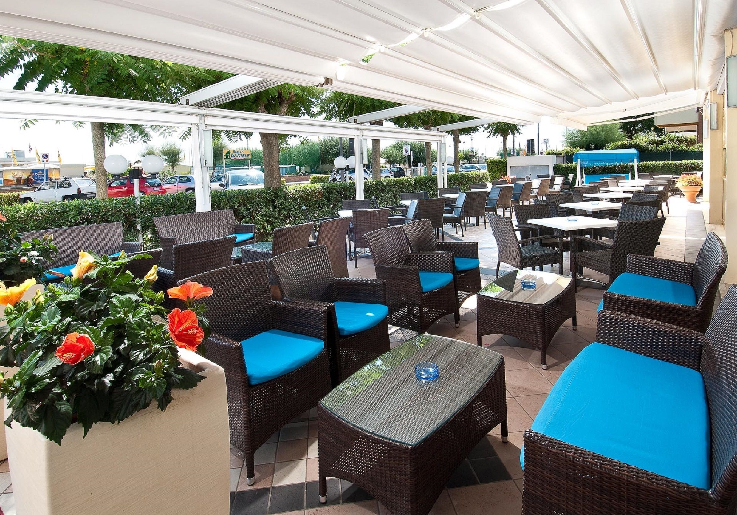 An outdoor cafe with wicker furniture and blue cushions, surrounded by greenery and a covered area.