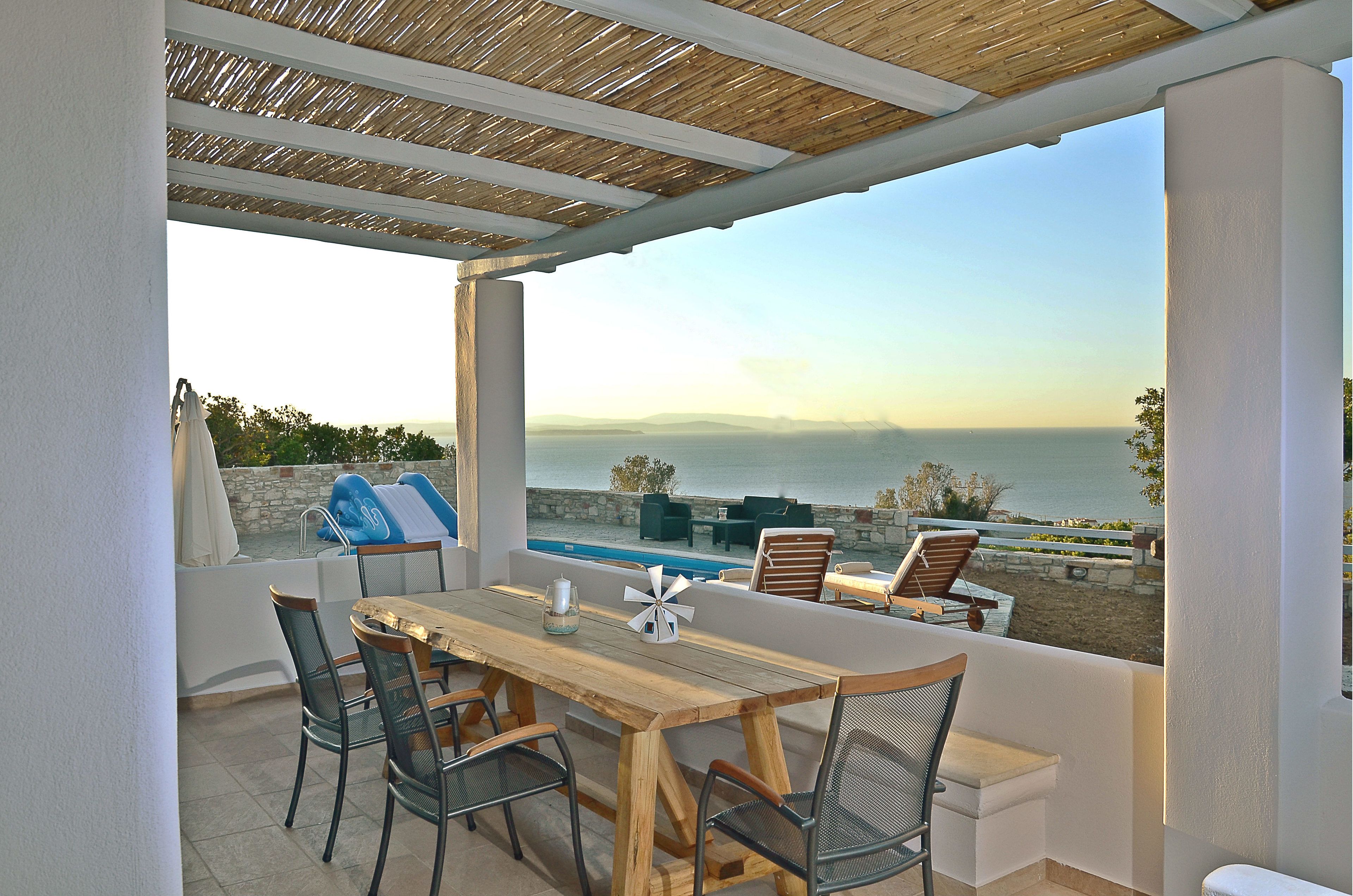 A covered outdoor seating area with a table and chairs, overlooking a pool and the sea.