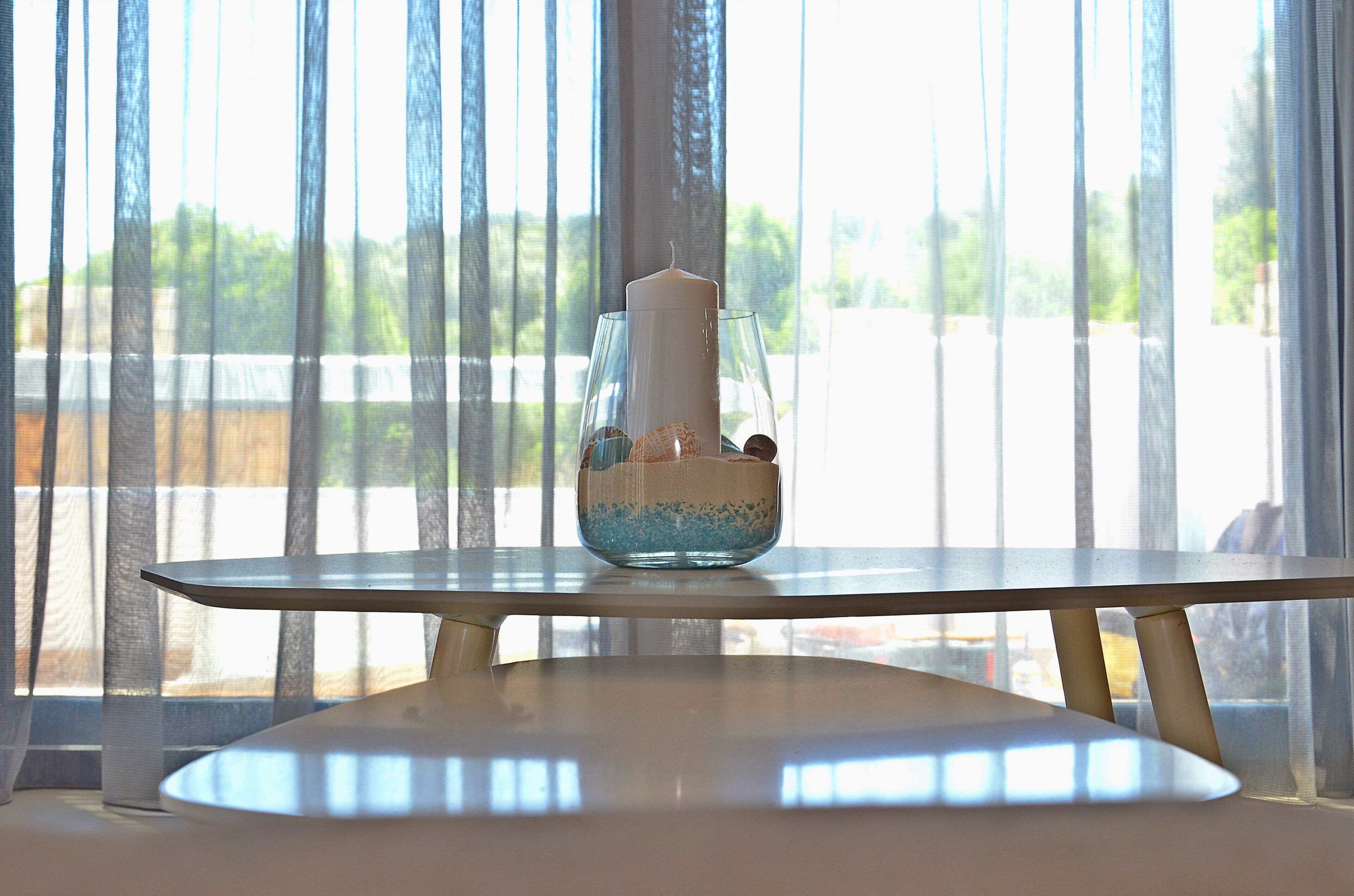 A table with a candle in a glass holder, a bowl with decorative items, and a view through sheer curtains.