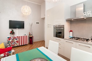 Una cocina moderna con estufa a gas, horno y microondas. Hay una zona de comedor con mesa y sillas. Un televisor está montado en la pared.