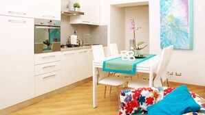 A modern kitchen with white cabinets, a built-in oven, and a sink. There is a dining table with chairs and a blue table runner. A colorful floral cushion is on the floor. - Bye Bye Roma Pantheon Suite (Rome)