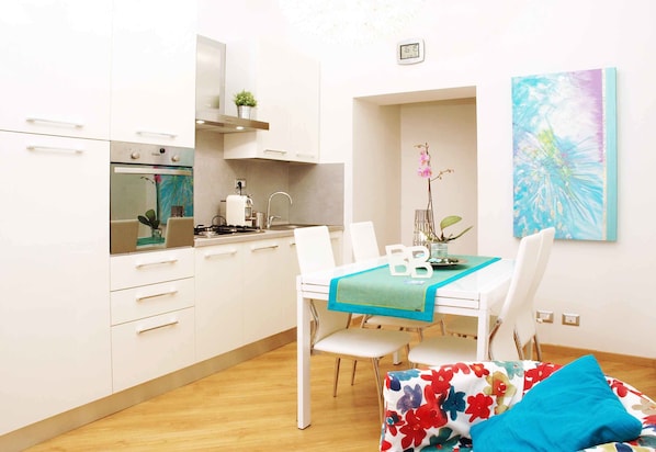 Una cocina moderna con armarios blancos, un horno empotrado y un lavabo. Hay una mesa de comedor con sillas y un camino de mesa azul. Un cojín floral y colorido está en el suelo.