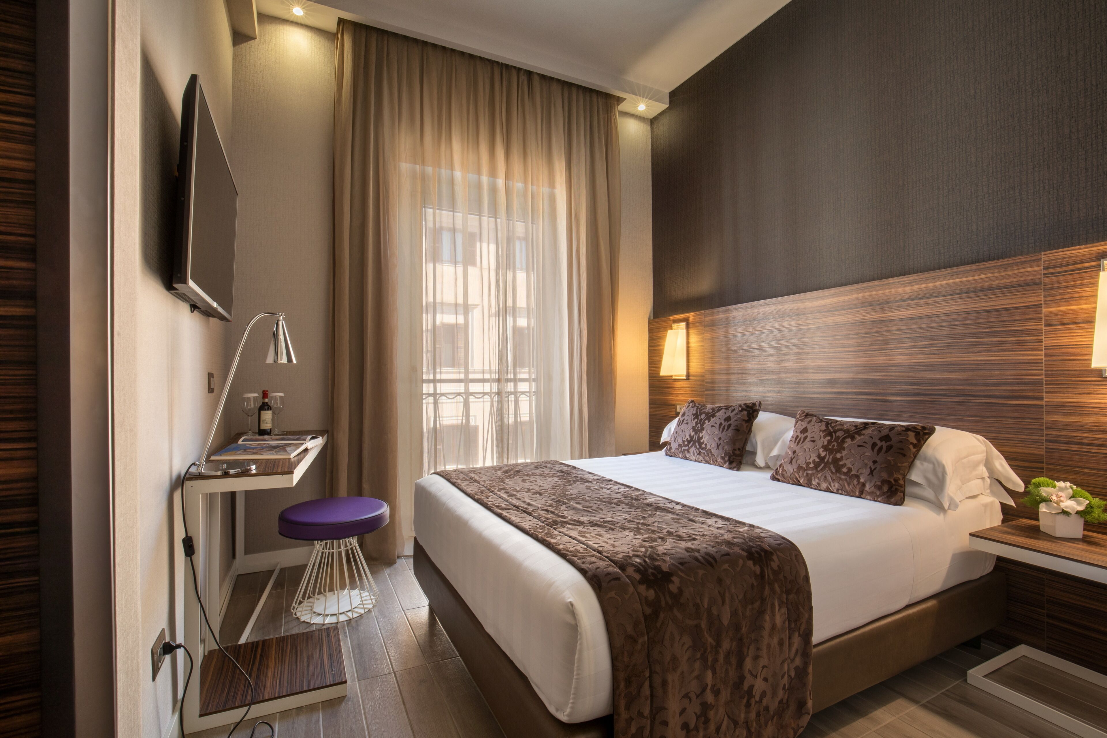 Une chambre d’hôtel moderne dotée d’un grand lit, d’un bureau avec une chaise, d’un téléviseur à écran plat fixé au mur et d’une fenêtre avec des rideaux.