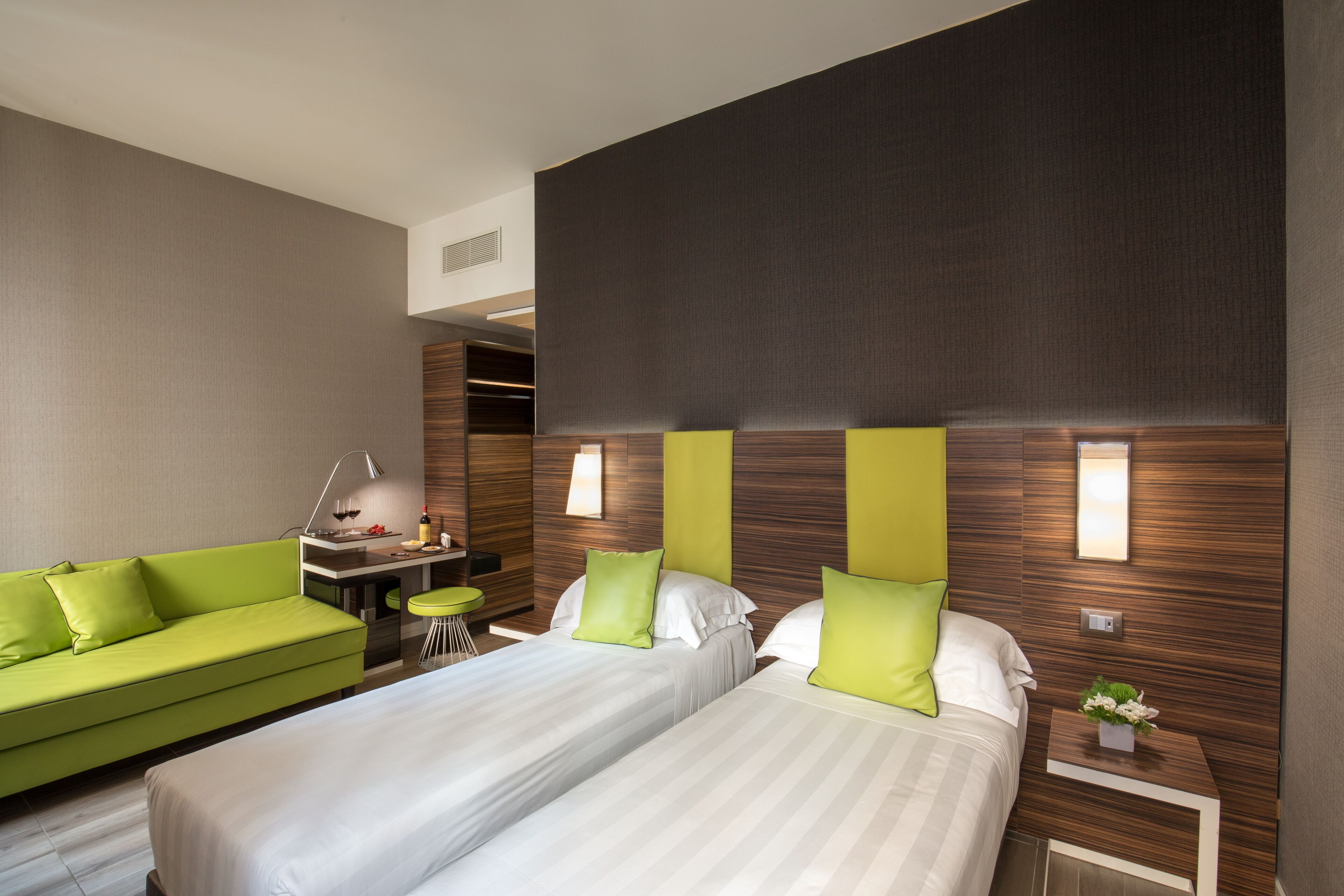 Une chambre d’hôtel avec deux lits, un canapé vert, un petit bureau avec une lampe et un luminaire fixé au mur.