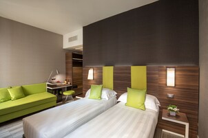 A hotel room with two beds, a green sofa, a small desk with a lamp, and a wall-mounted light fixture.