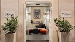 A hotel lobby with a reception desk, a seating area with a sofa and ottoman, and two potted plants. - The Republic Hotel (Rome)