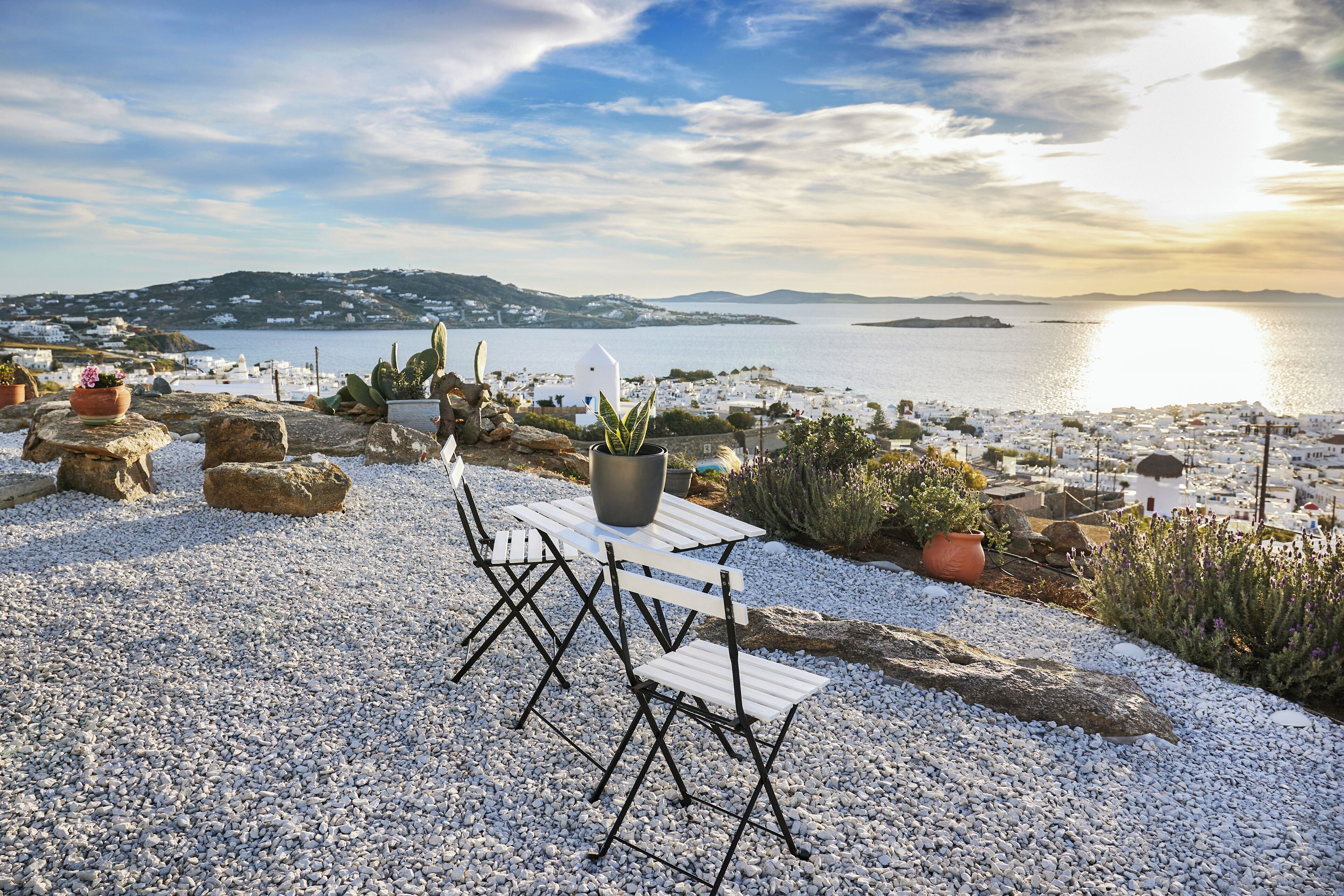 A terrace with white furniture, potted plants, and a view of a coastal town and sea.