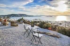 A terrace with white furniture, potted plants, and a view of a coastal town and sea. - Sand Lily Mykonos (Mykonos)