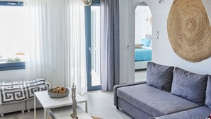 A modern living room with a white sofa, a grey couch, a white coffee table, and a woven rug. There is a ceiling fan, a woven wall hanging, and a view through an open door to a balcony. - Sand Lily Mykonos (Mykonos)