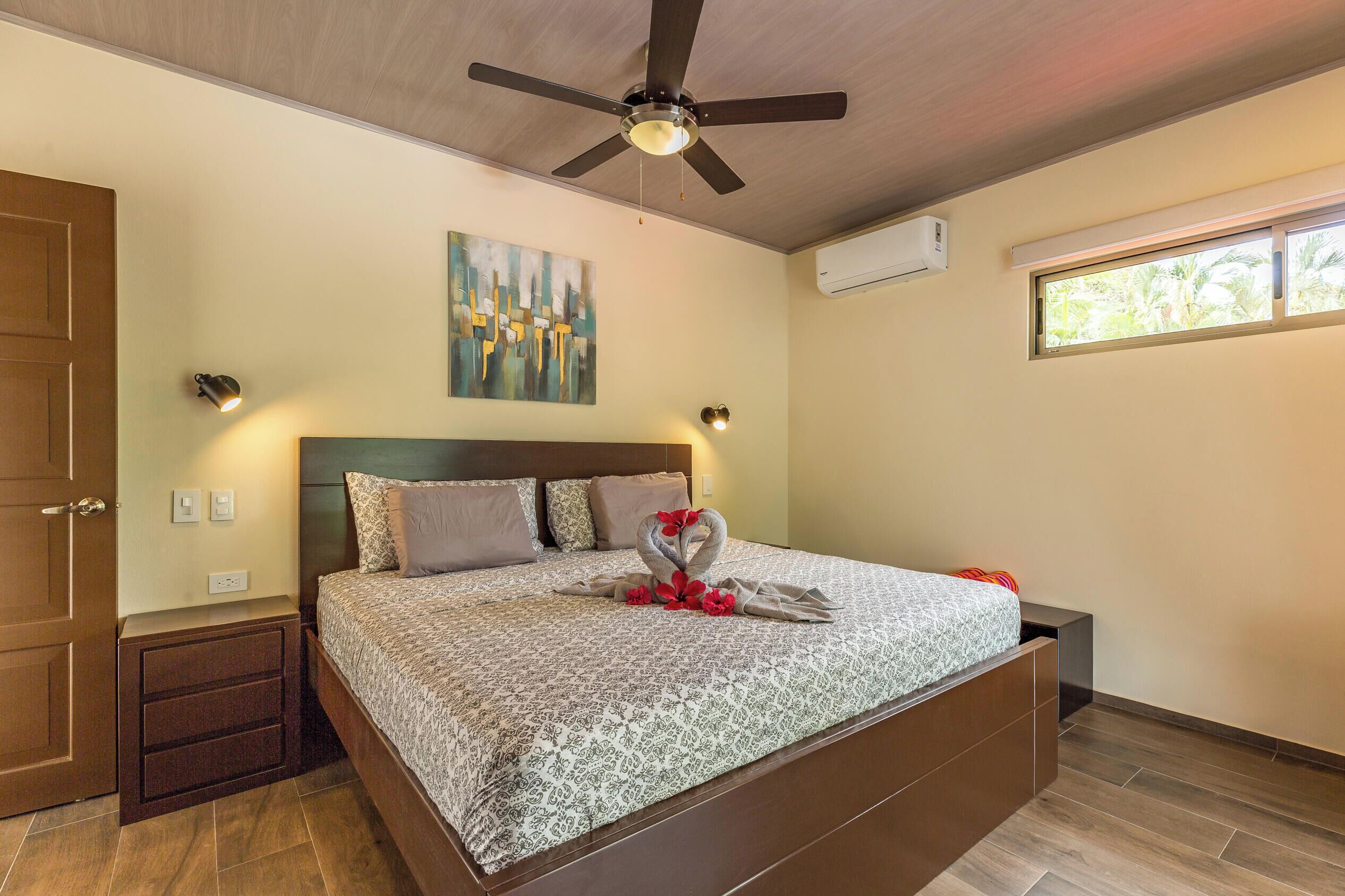 A bedroom with a bed, a ceiling fan, an air conditioning unit, a window, and a painting on the wall.